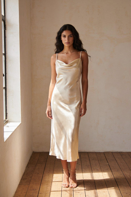 Woman wearing the Alma champagne satin bias-cut slip midi dress with a cowl neckline, photographed in natural daylight